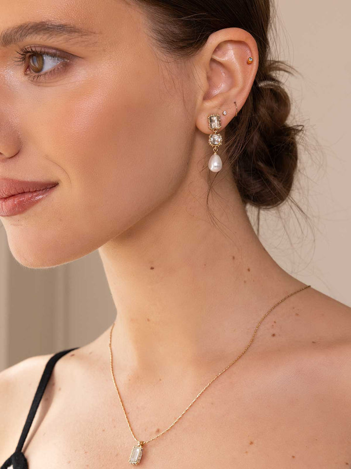 Close-up of a woman wearing a gold necklace and pearl earring against a neutral background